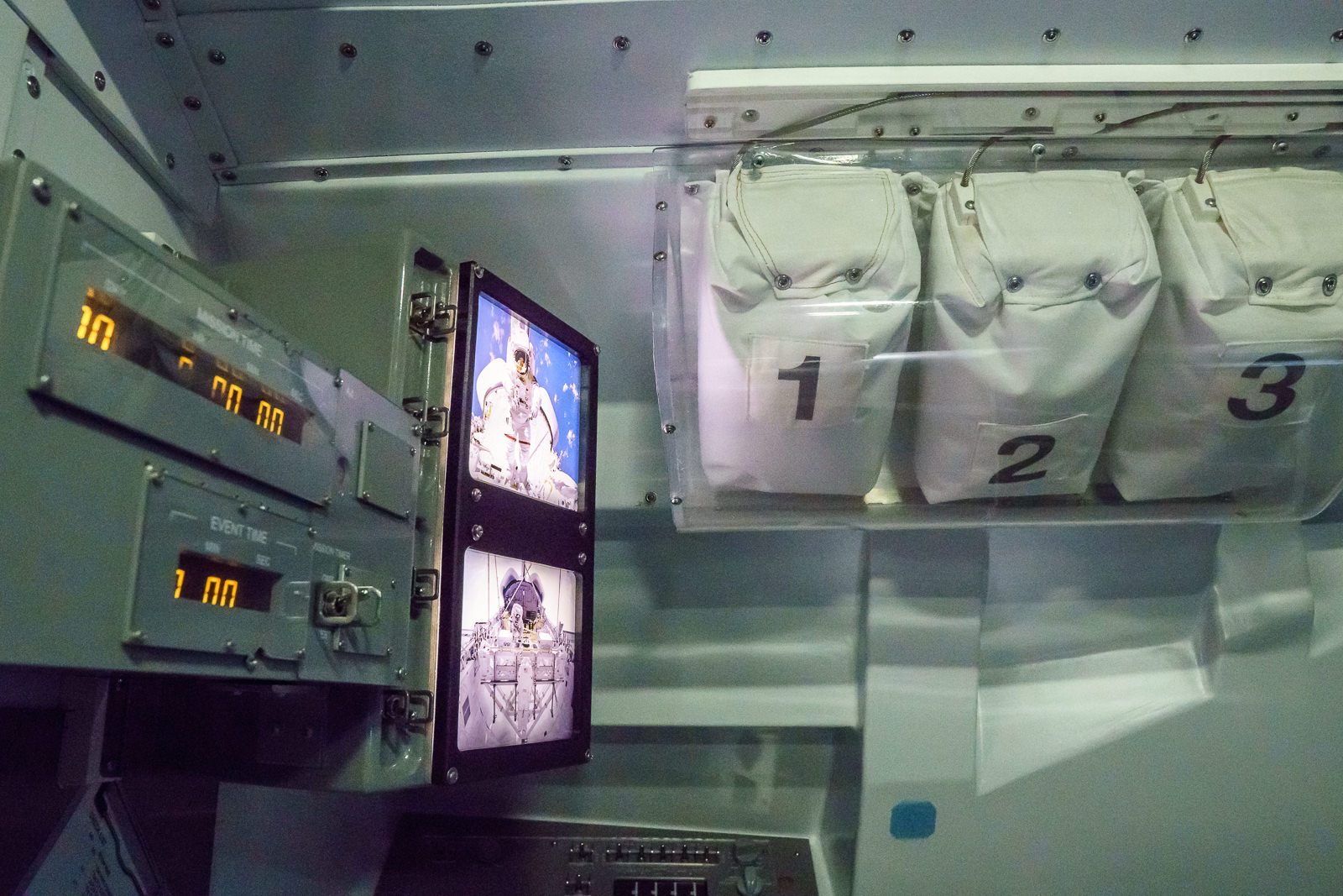 Telas do simulador de caminhada espacial mostrando cronômetro de missão e bolsas de equipamento numeradas