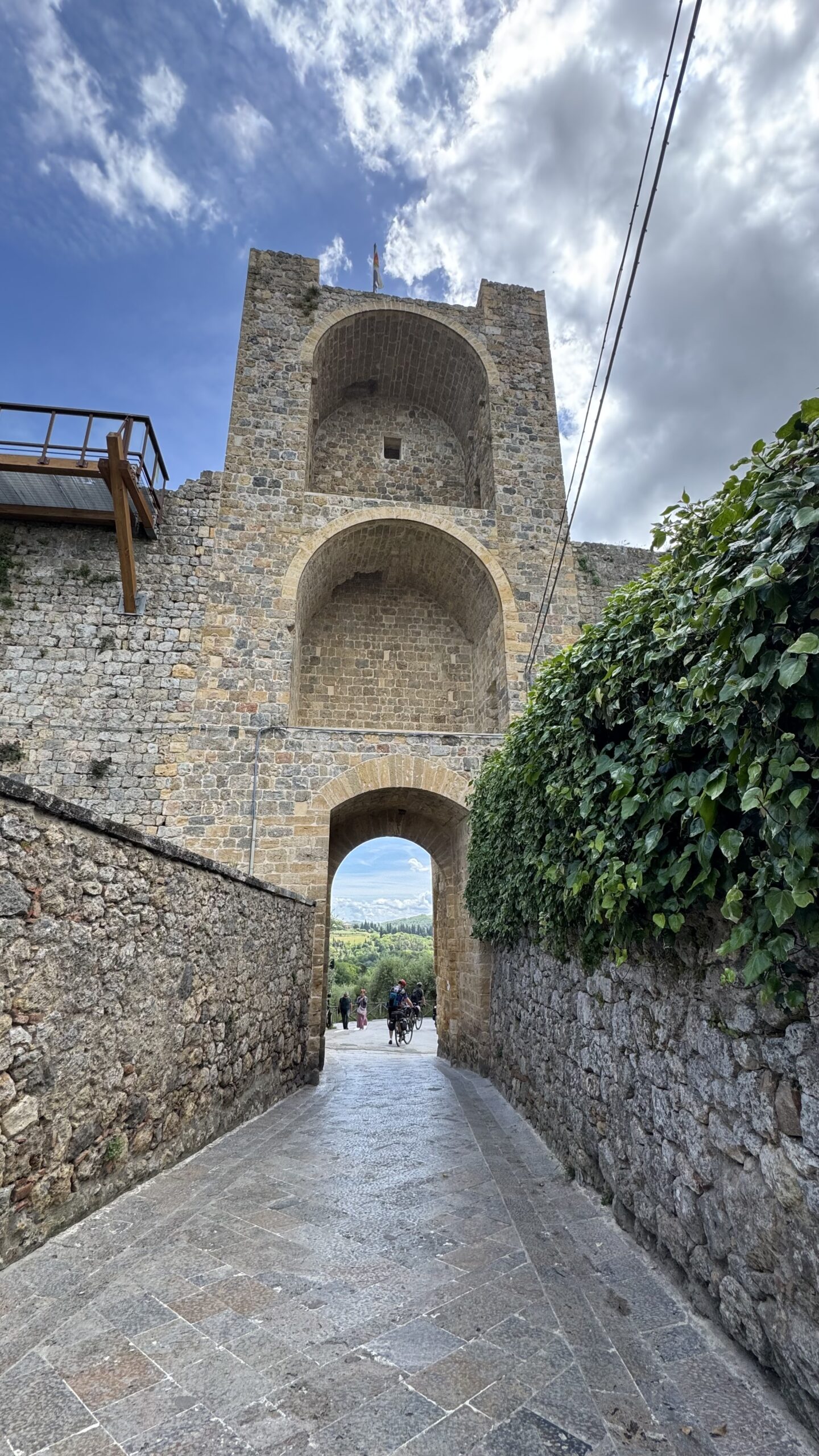 Vista de baixo do portão fortificado de Monteriggioni, com arcos de pedra em dois níveis e trecho de muralha coberto por hera