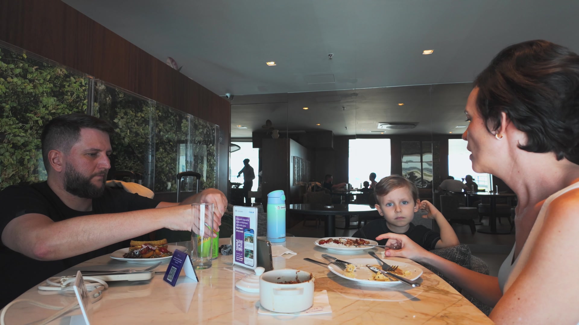 Rudi, Carol e o Sam à mesa fazendo uma refeição no Nubank Ultravioleta Lounge, em Guarulhos